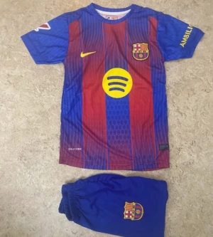 FC Barcelona Lamine Yamal matchtröja - Säljer en FC Barcelona matchtröja med Lamine Yamal och nummer 10 på ryggen. Tröjan är kortärmad, har klassiska blå och röda ränder, gula detaljer och klubbmärke på bröstet. Materialet är lätt polyester, perfekt för fotboll. Ingår matchande blå shorts med klubbmärke.