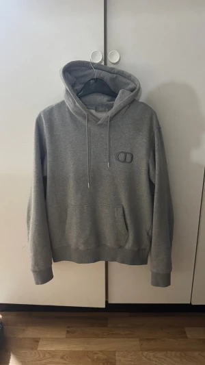 CD HOODIE - Snygg grå hoodie från CD med broderad CD-logga på bröstet. Tröjan har huva med snörning, ribbade muddar och en stor magficka. Tillverkad i mjuk bomullsmix som känns skön mot huden. Perfekt för dig som gillar stilrena och exklusiva plagg.