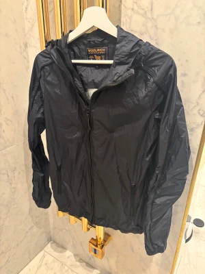 Svart vindjacka från Woolrich - Säljer en snygg svart vindjacka från Woolrich med huva och dragkedja framtill. Jackan har två fickor med dragkedja och är tillverkad i lätt polyester, perfekt för blåsiga dagar. Anpassbar huva och midja med spännen. Skönt och tunt material. Storlek på plagget S men realistiskt mer XS eller väldigt liten S i såna fall! Riktigt bra skick och inga defekter hittade! Pris: 1299kr 