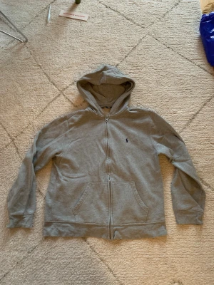 Grå hoodie från Polo Ralph Lauren  - Säljer en grå hoodie från Polo Ralph Lauren i storlek XL. Tröjan har dragkedja, huva och två fickor framtill. Klassisk broderad logga på bröstet och mjukt bomullsmaterial. Perfekt för chill dagar eller när du vill ha en clean look.