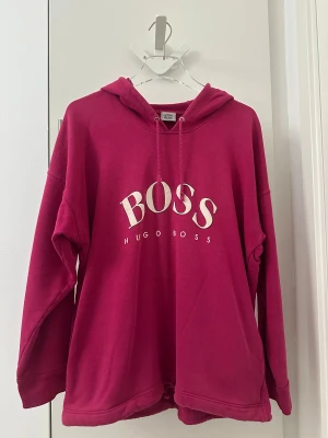 Hugo Boss Mörkrosa hoodie Large - Säljer en mörkrosa hoodie från Hugo Boss med stor vit logga på bröstet. Tröjan har huva med snörning, långa ärmar och praktiska fickor på sidorna. Nedtill finns justerbar snörning för extra passform. Materialet är mjuk bomull, perfekt för chill dagar.