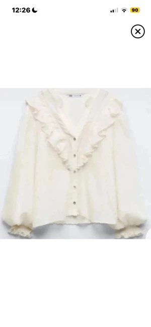 Vit blus med volanger från Zara - Säljer en vit blus från Zara med transparent prickigt tyg och stora volanger längs v-ringningen. Blusen har knappar framtill och puffiga långärmade ärmar med volangdetaljer vid mudden. Perfekt för dig som gillar romantisk och trendig stil.