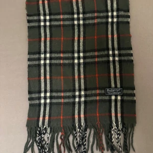 Rutig halsduk från Burberrys i ull - Snygg rutig halsduk från Burberrys i 100% lammull. Färgerna är grå, svart, vit och röd med klassiskt rutmönster och fransar i ändarna. Perfekt accessoar för att lyfta din stil under kyliga dagar. halsduken skickad från Norge och moms kan tilkomma. betalning måste ske via paypal eller liknande.