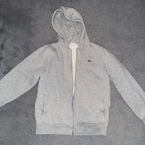 Lacoste kofta  - Snygg ljusgrå hoodie från Lacoste med klassisk krokodillogga på bröstet. Tröjan har huva med snörning, dragkedja framtill och två fickor. Tillverkad i mjuk bomullsblandning, perfekt för chill dagar eller när du vill ha en clean look.