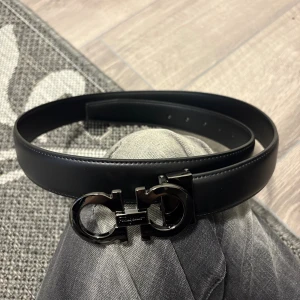 Svart skärp från Salvatore Ferragamo - Snyggt svart bälte från Salvatore Ferragamo i slätt skinn med ikoniskt dubbel-Gancini-spänne i blank svart metall. Bältet har en stilren och klassisk design som passar till jeans eller kostymbyxor. Perfekt accessoar för att lyfta din outfit.