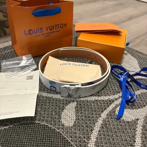 Louis Vuitton vitt skärp med monogram - Exklusivt vitt Louis Vuitton-bälte i skinn med ikoniskt blått och grått monogrammönster och silverfärgat LV-spänne. Bältet har en stilren design och levereras med originalpåse, box och kvitto. Perfekt accessoar för att lyfta din outfit.