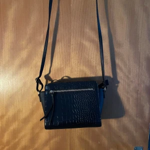 Svart axelväska med krokoprägling - Snygg svart axelväska med krokopräglad yta och justerbar rem. Väskan har en praktisk dragkedja framtill och stilren design som passar till många outfits. Perfekt storlek för det viktigaste och ger en cool vibe till din look.