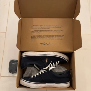 Mörkblå sneakers från Polo Ralph Lauren - Snygga mörkblå skor från Polo Ralph Lauren med klassisk vit sula och vita snören. Skorna har Polo-loggan broderad i guld på sidan och är tillverkade i slitstark bomull. Rund tå och platt sula ger en clean och sportig look. Perfekta för dig som gillar den trendiga klädstilen!