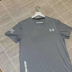 Grå Under Armour t-shirt slim fit - Snygg grå t-shirt från Under Armour med slim fit passform. T-shirten har korta ärmar, rund hals och coola logodetaljer i holografisk stil på bröstet, ärmen och nedtill. Perfekt för träning eller chill dagar. Materialet är lätt polyester som andas. Passar mig perfekt som är 178cm