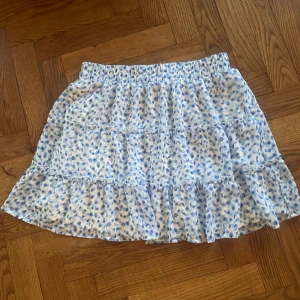 Blommig volangkjol från FB Sister - Supersöt kort kjol från FB Sister i vitt med blått blommönster. Kjolen har flera volanger och resår i midjan för skön passform. Tillverkad i lätt polyester som känns luftig och fräsch. Perfekt för dig som gillar romantisk och feminin stil.