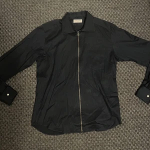 Svart skjorta med dragkedja John Henric - Svart skjorta från John Henric med klassisk krage och hel dragkedja framtill. Skjortan har långa ärmar med manschett och är tillverkad i bomull. Snygg och stilren design som sticker ut med sin zip-detalj.