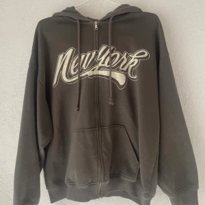 Mörkgrå hoodie med New York-tryck H&M - Säljer en mörkgrå hoodie från H&M Divided med stort vitt New York-tryck på bröstet. Tröjan har dragkedja, huva med snörning och känguruficka. Materialet är en mix av bomull och polyester, perfekt för chill dagar.