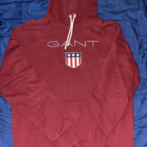 Vinröd hoodie från GANT  - Snygg vinröd hoodie från GANT i storlek XL med vit snörning och klassiskt tryck på bröstet med logga och amerikanskt sköldmotiv. Tröjan har huva, lång ärm och är tillverkad i mjuk bomull. Perfekt för chill dagar och streetstyle.