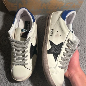 Golden Goose vita sneakers, skor - Golden Goose sneakers i vitt skinn med svart stjärna på sidan och blå häl. Endast andvända ett fåtal gånger och är i mycket bra skick. Pris kan självklart diskuteras vid snabb affär😃