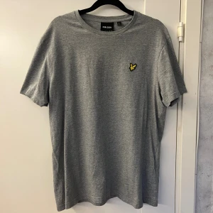 Grå t-shirt från Lyle & Scott - Snygg grå t-shirt från Lyle & Scott i storlek L. Klassisk rund hals och korta ärmar. På bröstet sitter den ikoniska gula fågel-loggan som ger en clean och stilren look. Tillverkad i mjuk bomull som känns skön mot huden. Den har inga tecken på användning så den är i fint skick. Hör av er om ni har frågor!