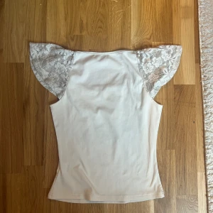 Vit topp med spetsärmar - Säljer en ljusvit topp med ribbad struktur och korta, volangiga spetsärmar med blommönster. Toppen har en mjuk, rundad halsringning och är figurnära i passformen. Perfekt för dig som gillar romantiska detaljer och en clean look.