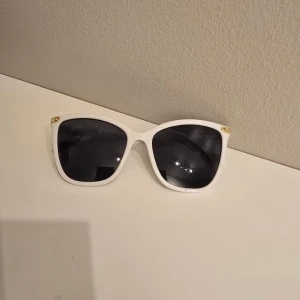 Vita solglasögon med svarta glas - Snygga solglasögon med vit båge och svarta glas. Klassisk fyrkantig form och små gulddetaljer vid gångjärnen. Perfekta för att lyfta din stil under soliga dagar.