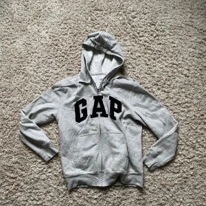Grå hoodie från GAP med dragkedja - Säljer en grå hoodie från GAP i storlek S med svart GAP-tryck på bröstet. Tröjan har huva med snörning, två fickor fram och dragkedja. Mjuk insida i fleece och klassisk loose passform. 