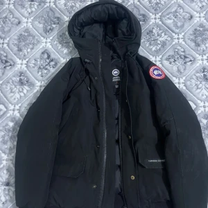 Svart dunjacka från Canada Goose - Svart dunjacka från Canada Goose med huva och klassisk logopatch på ärmen. Jackan har dragkedja och knappar framtill, samt praktiska fickor. Perfekt för kalla dagar och har en clean, stilren look som funkar till det mesta.