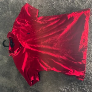 Röd Polo Ralph Lauren piké - Snygg röd pikétröja från Polo Ralph Lauren med klassisk krage och två knappar. Custom slim fit-modell med korta ärmar och ikonisk blå broderad logga på bröstet. Tillverkad i mjuk bomull som är skön mot huden. Perfekt för en chill och stilren look.