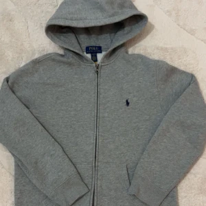 Grå hoodie från Polo Ralph Lauren - Snygg grå hoodie från Polo Ralph Lauren med klassisk blå broderad logga på bröstet. Tröjan har huva, hel dragkedja och två fickor framtill. Perfekt för chill dagar och enkel att matcha med jeans eller joggers.