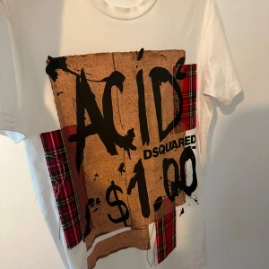 Vit DSquared t-shirt med tryck och rutor - Cool vit t-shirt från DSquared med stor grafisk print 'ACID $1.00' i svart och brunt framtill. T-shirten har röda och svarta rutiga tygdetaljer som ger en edgy vibe. Klassisk rund hals och korta ärmar. Perfekt för dig som gillar statement-plagg. Använd 2 gånger 