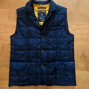 Mörkblå quiltad dunväst Oscar Jacobson - Quiltad mörkblå dunväst från Oscar Jacobson med gul insida. Västen har tre framfickor, knappar och klassisk krage. Perfekt för lager-på-lager och har en stilren, modern look. Materialet är glansig polyester och dunfyllning.