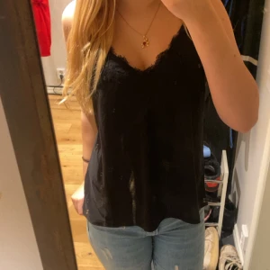 Svart linnetopp med spetskant - Svart linnetopp med smala axelband och v-ringning dekorerad med spetskant. Tunt och lätt material som ger en luftig känsla. Perfekt att matcha med jeans eller kjol för en stilren look.