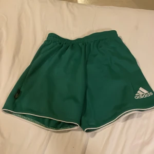 Gröna Adidas shorts med vit kant - Snygga gröna shorts från Adidas med vit kant och broderad logga på benet. Shortsen har elastisk midja och innerbyxa i vitt meshmaterial, perfekta för sport eller bad. Klassisk sportig look och bekväm passform.