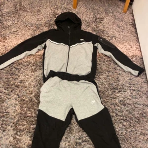 Nike tech - Nike hoodie i svart och grå med dragkedja och huva. Tröjan har lång ärm, vit detalj på bröstet och sportig design. Perfekt för träning eller chill. Matchande byxor ingår ej.