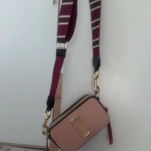 Marc Jacobs Snapshot axelväska rosa - Snygg Marc Jacobs Snapshot axelväska i ljusrosa och beige med guldfärgade detaljer. Väskan har ett brett, randigt axelband i vinrött och beige, samt dubbla dragkedjor och det ikoniska Marc Jacobs-logot på framsidan. Perfekt storlek för mobilen och småsaker.