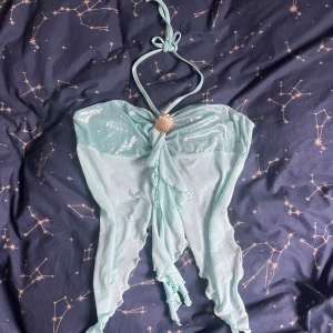 Blå topp med snäckdetalj - Säljer en unik blå topp med glittrigt tyg upptill och transparent mesh nedtill. Toppen har halterneck med knyt och en snygg snäckformad detalj framtill. Volangliknande kanter ger en extra cool look, perfekt för stranden eller poolen. Använd en gång på halloween i år bara och säljer för den är out of my comfort zone.
