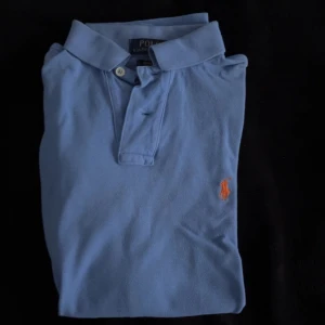Blå pikéskjorta från Polo Ralph Lauren - Snygg ljusblå pikéskjorta från Polo Ralph Lauren i storlek S, custom fit. Klassisk krage, korta ärmar och två knappar framtill. Ikonisk orange logga broderad på bröstet. Skjortan är gjord i mjuk bomull som känns skön mot huden. Hör av dig om du har några frågor.