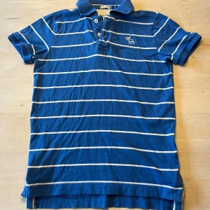 Blå randig piké från Abercrombie & Fitch - Snygg blå polo från Abercrombie & Fitch i storlek S, med vita horisontella ränder och klassisk krage. Tröjan har korta ärmar, knappar framtill och en broderad älg på bröstet. Tillverkad i mjuk bomull, perfekt för en chill och stilren look.