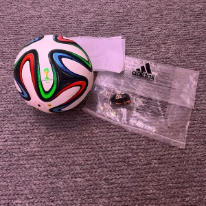 Adidas Brazuca VM 2014 fotboll - Säljer en Adidas Brazuca officiell matchboll från VM 2014 i Brasilien. Bollen är vit med färgstarka detaljer i blått, grönt, rött och svart. FIFA World Cup-logga tryckt på sidan. Tillverkad i slitstarkt syntetmaterial och levereras med originalpåse. Går inte att få tag på nya. Helt oanvänd till bra pris. Kan gå ner i pris vid snabb affär 