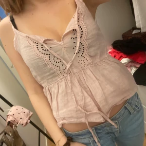 Ljusrosa ärmlös linnetopp med broderi - Supersöt ljusrosa linnetopp med tunna axelband och broderade detaljer framtill. Toppen har knytning vid bysten och en lätt volang nedtill som ger en avslappnad och feminin look. Perfekt för varma dagar och enkel att matcha med jeans eller kjol.