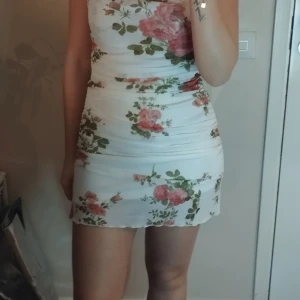 Vit blommig kortklänning med smala axelband - Supersöt vit kortklänning med smala justerbara axelband och blommigt mönster i rosa och grönt. Klänningen har en figurnära passform och lätt rynkade detaljer längs sidorna för en snygg siluett. Perfekt för dig som gillar romantisk och feminin stil.