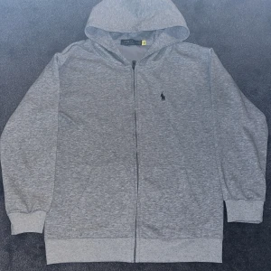 Grå hoodie från Polo Ralph Lauren - Grå hoodie från Polo Ralph Lauren med klassisk svart logobrodyr på bröstet. Tröjan har huva, hel dragkedja och två fickor framtill. Tillverkad i mjuk bomullsmix med ribbade muddar och lång ärm. Perfekt för chill dagar och streetwear.