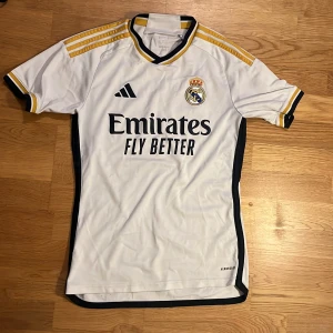 Real Madrid vit matchtröja Adidas - Snygg Real Madrid fotbollströja från Adidas i vit med svarta och gula detaljer, perfekt för träning eller match. Kopte den i spanien 1500kr