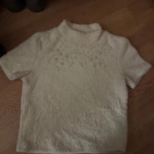 Vit fluffig tröja med pärlor från Zara - Superfin vit fluffig tröja från Zara med korta ärmar och hög krage. Tröjan har pärldetaljer framtill som ger en extra touch. Materialet är mjukt och känns lyxigt, perfekt för dig som gillar att sticka ut med detaljer.