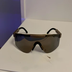 Guldiga Pit Viper sportglasögon - Säljer ett par feta Pit Viper solglasögon med guldiga detaljer och svarta bågar. De har en stor, speglande lins i shield-modell som ger maxad look och skydd. Perfekta för dig som vill sticka ut och köra på en sportig vibe.