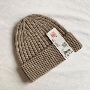 Beige ribbstickad mössa från Uniqlo - Snygg beige ribbstickad mössa från Uniqlo HEATTECH. Klassisk modell med uppvikt kant och grovstickad struktur som håller dig varm. Perfekt accessoar för kyliga dagar och enkel att matcha med alla outfits.