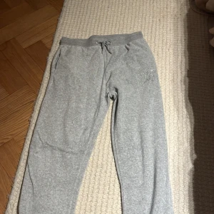 Grå mjukisbyxor Juicy Couture - Grå mjukisbyxor från Juicy Couture med glittrande JC-logo i strass på ena höften. Byxorna har snörning i midjan, två fickor bak och är tillverkade i mjukt bomullsmaterial. Perfekta för en chill och avslappnad stil.