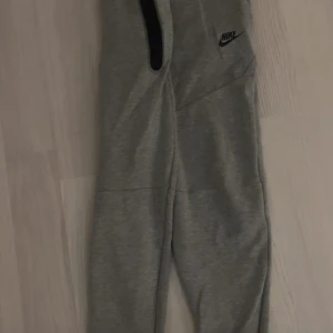 Grå mjukisbyxor från Nike med snörning - Grå mjukisbyxor från Nike med svart logga på vänster ben, svarta snören i midjan och svarta detaljer vid fickorna. Byxorna har normal passform och är tillverkade i mjukt bomullsmaterial. Perfekta för chill eller träning.