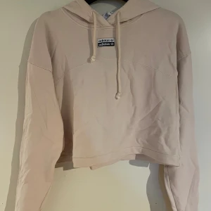 Ljusbeige croppad hoodie från Adidas - Säljer en ljusbeige croppad hoodie från Adidas med broderad logga fram och tryck bak. Tröjan har huva med snörning och långa ärmar. Tillverkad i mjuk bomull, perfekt för chill dagar. Adidas Originals-märke både fram och bak för extra streetstyle.