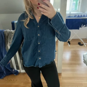 Blå jeansskjorta med knappar - Snygg blå jeansskjorta med guldiga knappar och två bröstfickor. Skjortan har lång ärm och klassisk krage, perfekt att styla till olika outfits. Materialet är mjukt och ger en avslappnad känsla.