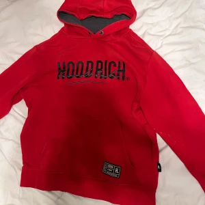 Röd hoodie från Hoodrich i storlek L - Snygg röd hoodie från Hoodrich med svart logotyp tryckt både fram och bak. Tröjan har en stor magficka, huva med grått foder och coola detaljer som tryck vid nederkanten. Perfekt för dig som gillar streetwear och vill sticka ut. Lite små fläker på armen med de går att få bort