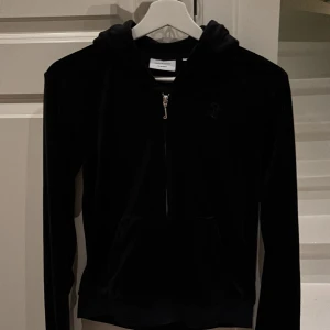 Svart velour hoodie Juicy Couture - Svart hoodie från Juicy Couture i mjuk velour med dragkedja framtill och broderad JC-logga på bröstet. Klassisk huva och känguruficka, perfekt för en chill och trendig look.