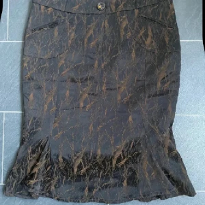 Svart och brun mönstrad kjol Favori - Unik svart kjol med brunt abstrakt mönster från Favori. Kjolen har snygga detaljer som dekorativa knappar, fickor framtill och plisserad nederkant bak. Passformen är normal och materialet känns som polyester. Perfekt för dig som gillar coola detaljer och mönster.