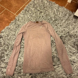 Rosa långärmad träningströja Gymshark - Superskön långärmad träningströja från Gymshark i ljusrosa nyans. Tumhål vid ärmslut och diskret mönster i tyget. Perfekt för gymmet eller löprundan, med snyggt logotryck på bröstet. 